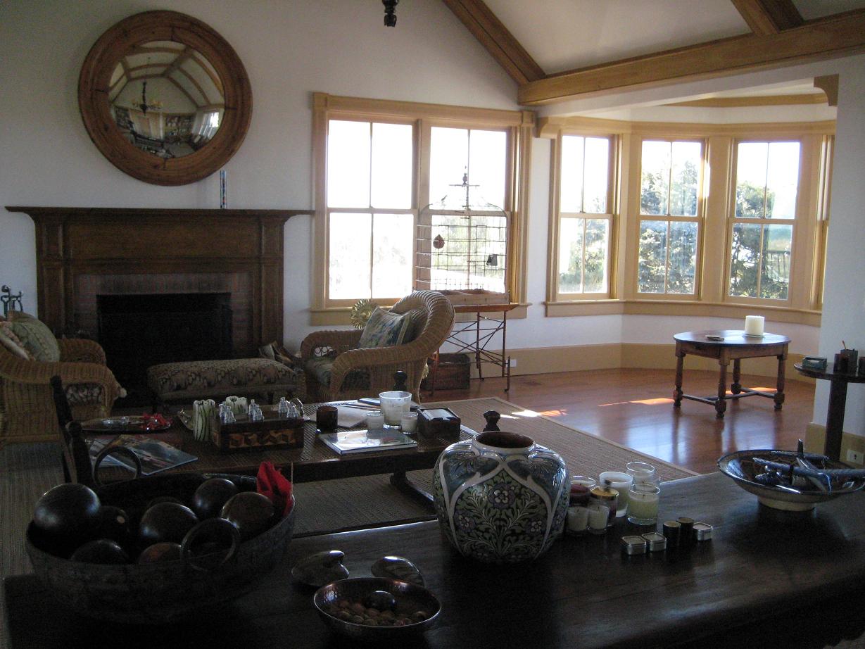 Jasne wnętrze salonu z drewnianymi oknami, kominkiem z lustrem i wiklinowymi fotelami, oświetlone światłem słonecznym.